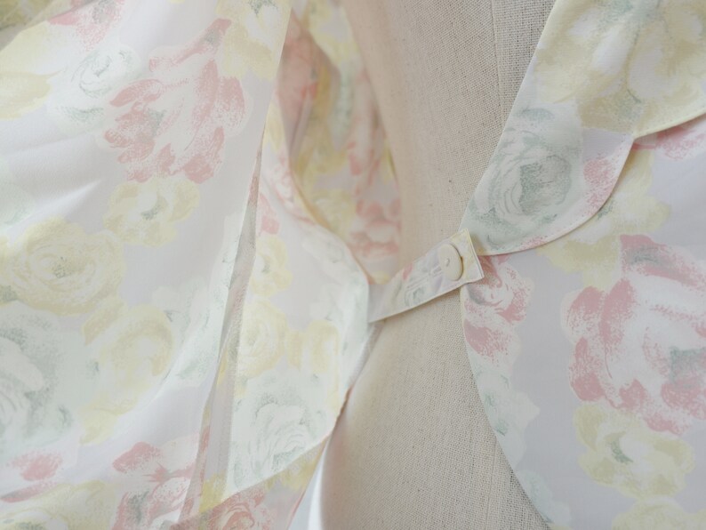 May include: A white blouse with a floral print featuring pink, yellow, and green roses. The blouse has a large, rounded collar with a button closure.