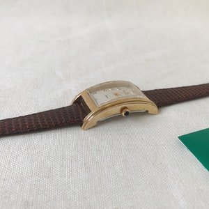 K&ouml;nnte beinhalten: Eine goldfarbene Armbanduhr mit wei&szlig;em Zifferblatt und braunem Lederarmband. Die Uhr hat eine rechteckige Form und ein klassisches Design.