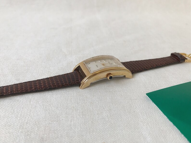 K&ouml;nnte beinhalten: Eine goldfarbene Armbanduhr mit wei&szlig;em Zifferblatt und braunem Lederarmband. Die Uhr hat eine rechteckige Form und ein klassisches Design.