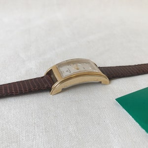K&ouml;nnte beinhalten: Eine goldfarbene Armbanduhr mit einem braunen Lederarmband. Das Zifferblatt der Uhr ist rechteckig und hat einen wei&szlig;en Hintergrund mit schwarzen r&ouml;mischen Ziffern als Stundenmarkierungen.
