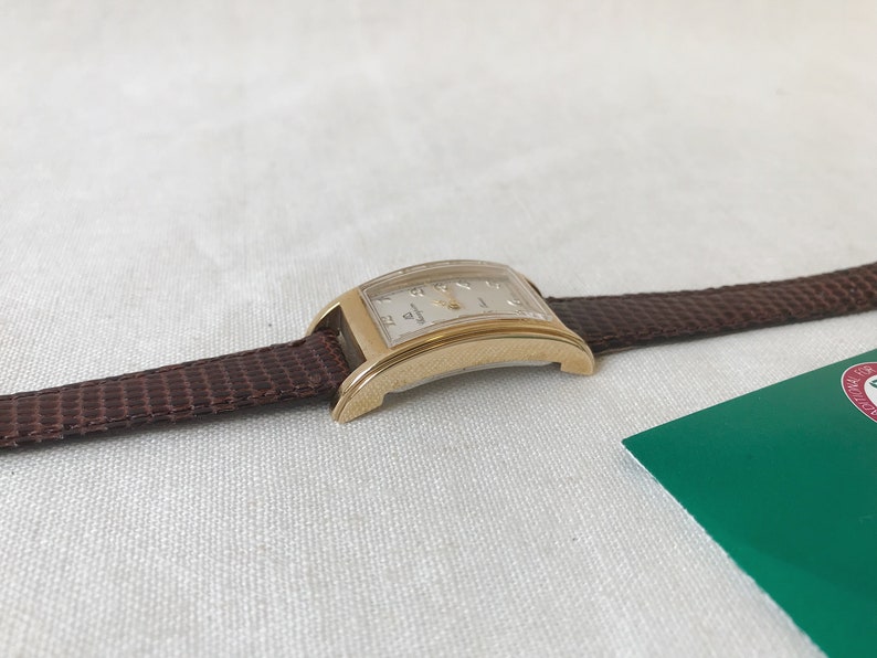 K&ouml;nnte beinhalten: Eine goldfarbene Armbanduhr mit einem braunen Lederarmband. Das Zifferblatt der Uhr ist rechteckig und hat einen wei&szlig;en Hintergrund mit schwarzen r&ouml;mischen Ziffern als Stundenmarkierungen.