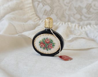 May include: A small black glass perfume bottle with a gold top and a white oval label with embroidered red roses. The bottle is sitting on a white fabric surface.