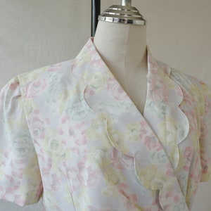 May include: A white blouse with a floral print featuring pink, yellow, and green flowers. The blouse has a scalloped collar and short sleeves.