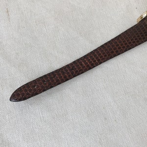 K&ouml;nnte beinhalten: Ein braunes Lederarmband mit einem goldenen Zifferblatt. Das Zifferblatt hat schwarze r&ouml;mische Ziffern als Stundenmarkierungen und einen wei&szlig;en Hintergrund.