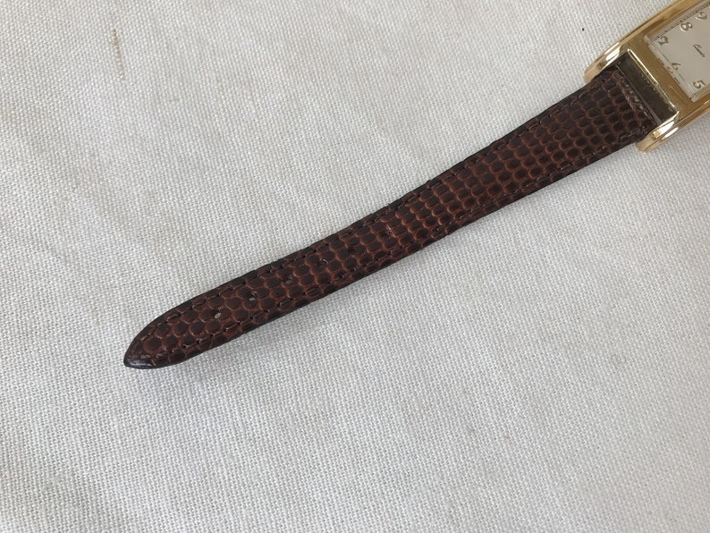 K&ouml;nnte beinhalten: Ein braunes Lederarmband mit einem goldenen Zifferblatt. Das Zifferblatt hat schwarze r&ouml;mische Ziffern als Stundenmarkierungen und einen wei&szlig;en Hintergrund.