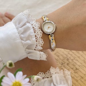 Può includere: Un orologio da polso argentato e dorato con un quadrante bianco e un design delicato e intricato. L'orologio è indossato su un polso con una blusa bianca con volant.