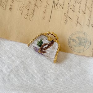 Puede incluir: Un pequeño bolso de mano blanco bordado con un marco dorado y un diseño floral. El bolso de mano es un broche vintage.