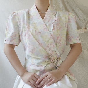 May include: A white blouse with a floral print and a scalloped collar. The blouse has a wrap style and is cinched at the waist.