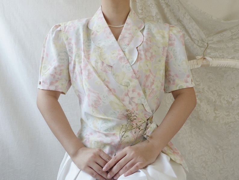 May include: A white blouse with a floral print and a scalloped collar. The blouse has a wrap style and is cinched at the waist.