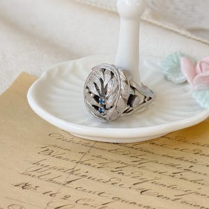 May include: A silver ring featuring a detailed leaf design and two blue gemstones. The ring is displayed on a white ring holder.