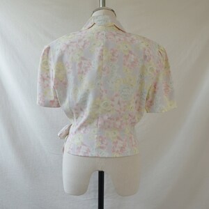 May include: A white blouse with a floral print in shades of pink, yellow, and green. The blouse has a wrap style and short sleeves.