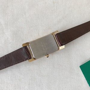 K&ouml;nnte beinhalten: Ein braunes Lederarmband mit einem rechteckigen goldfarbenen Uhrengeh&auml;use. Die R&uuml;ckseite des Uhrengeh&auml;uses ist silberfarben und tr&auml;gt den Aufdruck "WATER RESISTANT".