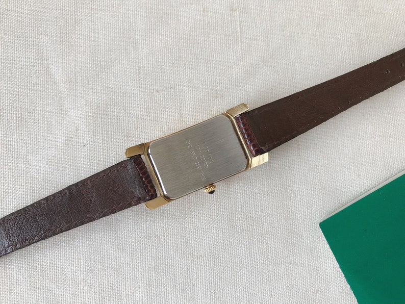 K&ouml;nnte beinhalten: Ein braunes Lederarmband mit einem rechteckigen goldfarbenen Uhrengeh&auml;use. Die R&uuml;ckseite des Uhrengeh&auml;uses ist silberfarben und tr&auml;gt den Aufdruck "WATER RESISTANT".