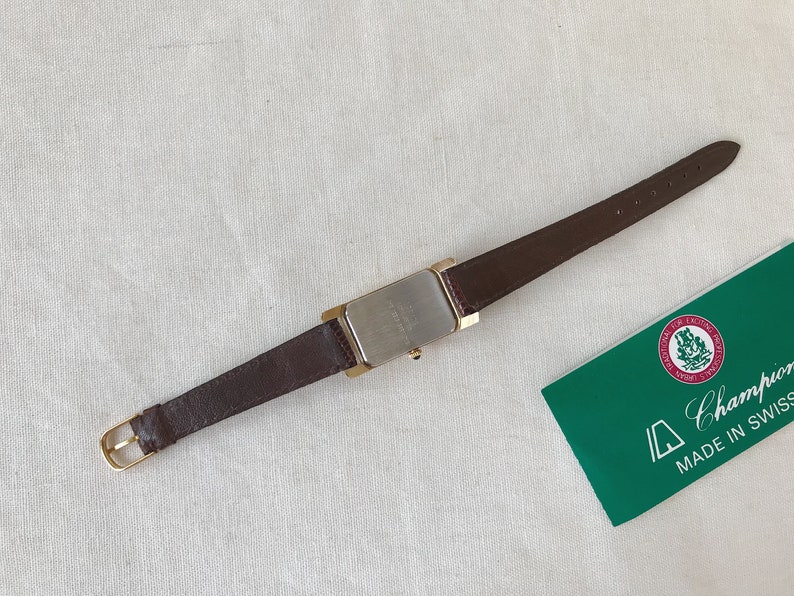 K&ouml;nnte beinhalten: Eine goldfarbene Armbanduhr mit einem braunen Lederarmband. Das Zifferblatt der Uhr ist rechteckig und hat eine silberfarbene Oberfl&auml;che. Die Uhr liegt auf einer wei&szlig;en Oberfl&auml;che. Die Uhr hat den Text "Champion Made in Swiss" auf einer gr&uuml;nen Karte.