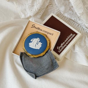 May include: A Stratton compact with a blue and gold design. The compact features a white cameo of a couple dancing. The box reads "Stratton compact" and "Stratton matching handbag accessories".