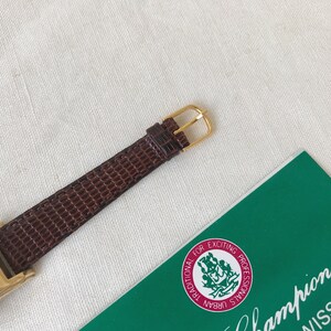 K&ouml;nnte beinhalten: Eine goldfarbene Uhr mit einem braunen Lederarmband. Das Zifferblatt der Uhr ist rechteckig und hat einen wei&szlig;en Hintergrund mit schwarzen r&ouml;mischen Ziffern als Stundenmarkierungen. Die Uhr liegt auf einer wei&szlig;en Oberfl&auml;che mit einer gr&uuml;nen Karte, auf der "Champion Swiss" und "Traditional for Exciting Professionals Urban Signals" steht.