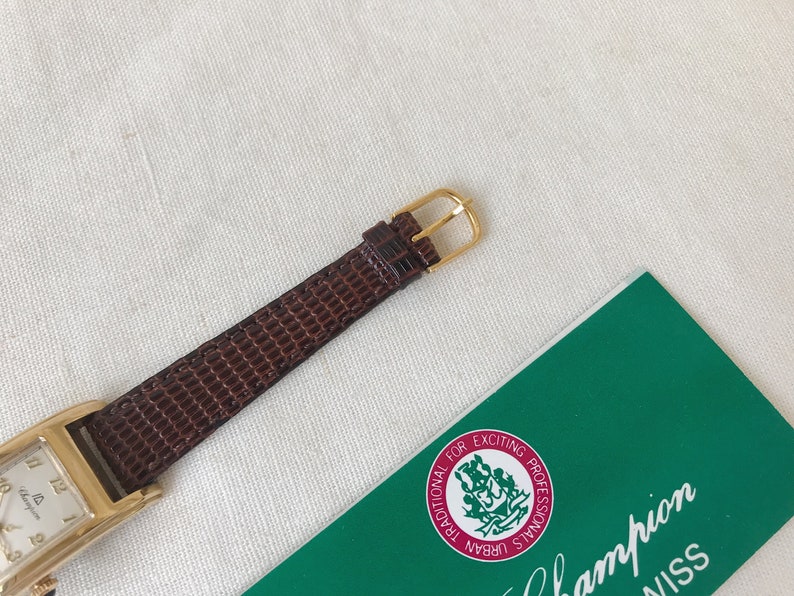 K&ouml;nnte beinhalten: Eine goldfarbene Uhr mit einem braunen Lederarmband. Das Zifferblatt der Uhr ist rechteckig und hat einen wei&szlig;en Hintergrund mit schwarzen r&ouml;mischen Ziffern als Stundenmarkierungen. Die Uhr liegt auf einer wei&szlig;en Oberfl&auml;che mit einer gr&uuml;nen Karte, auf der "Champion Swiss" und "Traditional for Exciting Professionals Urban Signals" steht.