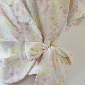 May include: A white blouse with a floral print in shades of pink, yellow, and green. The blouse has a tie at the waist and a ruffled collar.