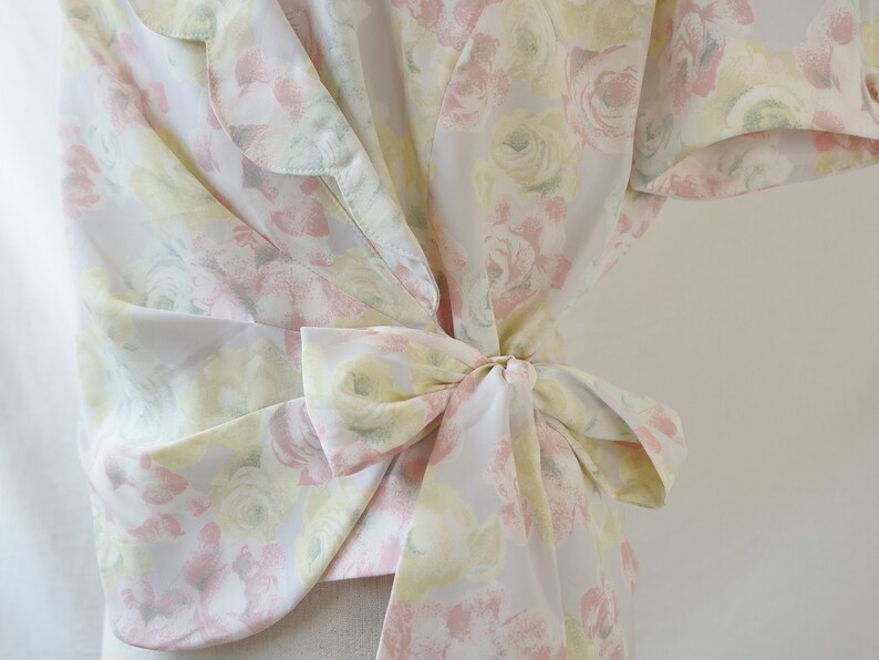 May include: A white blouse with a floral print in shades of pink, yellow, and green. The blouse has a tie at the waist and a ruffled collar.