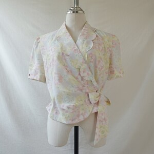 May include: A white, pink, and yellow floral print wrap blouse with a scalloped collar and short sleeves. The blouse has a tie at the waist.