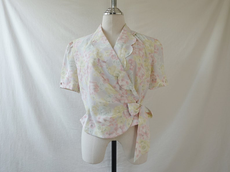 May include: A white, pink, and yellow floral print wrap blouse with a scalloped collar and short sleeves. The blouse has a tie at the waist.