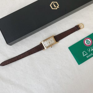 K&ouml;nnte beinhalten: Eine rechteckige Armbanduhr in Goldton mit wei&szlig;em Zifferblatt und braunem Lederband. Die Uhr befindet sich in einer Box mit einem Logo auf der Oberseite. Die Uhr wird au&szlig;erdem mit einer gr&uuml;nen Karte geliefert, auf der "Champion Made in Swiss" steht.