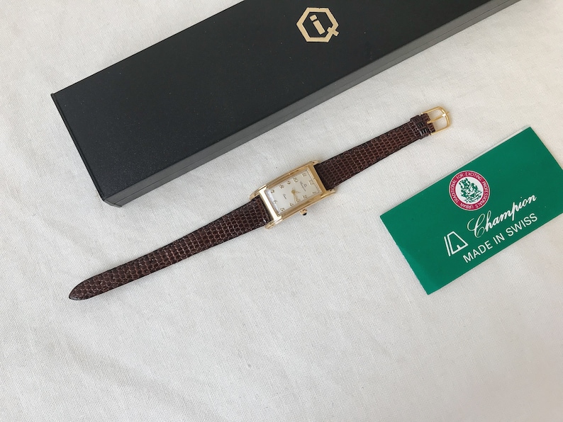 K&ouml;nnte beinhalten: Eine rechteckige Armbanduhr in Goldton mit wei&szlig;em Zifferblatt und braunem Lederband. Die Uhr befindet sich in einer Box mit einem Logo auf der Oberseite. Die Uhr wird au&szlig;erdem mit einer gr&uuml;nen Karte geliefert, auf der "Champion Made in Swiss" steht.