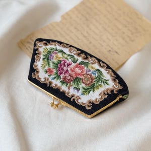 Könnte beinhalten: Eine schwarze Stofftasche mit einem goldenen Verschluss. Die Tasche ist mit einem floralen Stickmuster in Rosa-, Grün- und Weißtönen verziert.