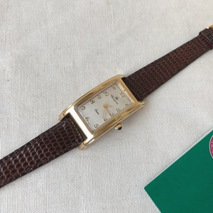 K&ouml;nnte beinhalten: Eine goldfarbene Uhr mit wei&szlig;em Zifferblatt und braunem Lederarmband. Die Uhr hat r&ouml;mische Ziffern als Stundenmarkierungen und ein Datumsfenster bei 3 Uhr.