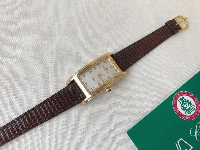 K&ouml;nnte beinhalten: Eine goldfarbene Uhr mit wei&szlig;em Zifferblatt und braunem Lederarmband. Die Uhr hat r&ouml;mische Ziffern als Stundenmarkierungen und ein Datumsfenster bei 3 Uhr.