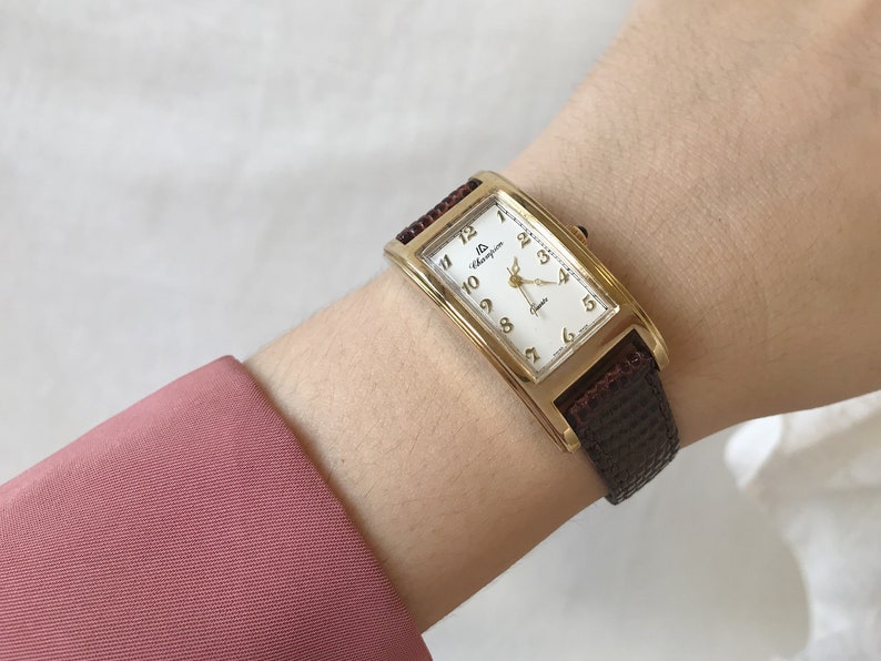 K&ouml;nnte beinhalten: Eine rechteckige goldfarbene Uhr mit wei&szlig;em Zifferblatt und braunem Lederarmband. Das Zifferblatt der Uhr hat schwarze r&ouml;mische Ziffern als Stundenmarkierungen und ein kleines Sekunden-Zifferblatt. Die Marke der Uhr ist "Champagne" und der Text "Automatic" befindet sich unter dem Zifferblatt.