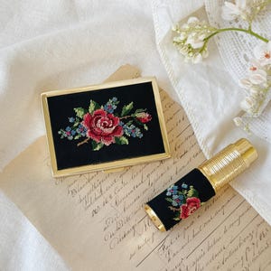 May include: A gold compact mirror with a black velvet interior featuring an embroidered floral design. The mirror is next to a gold and black lipstick tube with a similar floral design.