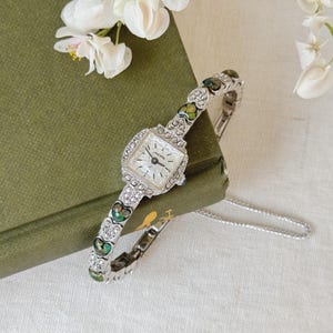 Puede incluir: Un reloj de pulsera plateado con una esfera cuadrada y una banda con estampado verde y plateado. El reloj está sobre un libro verde con flores blancas.