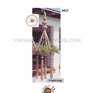 Puede incluir: Un macetero de macramé de 1,5 metros de largo, hecho de cuerda de fibra natural. El macetero tiene forma de triángulo con una parte superior decorativa y borlas en la parte inferior. El macetero está colgado de una puerta.