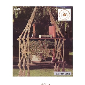 May include: A macrame plant hanger with two shelves, made of natural rope with wooden accents. The hanger is 5.5 feet long and features a decorative tassel at the bottom. The hanger is shown with a potted plant and decorative items on the shelves.