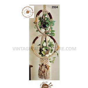 May include: A macrame plant hanger with two circular holders, each with a plant. The top ring has a dark brown accent, and the bottom ring has a lighter brown accent. The text "2934" is visible. The image also includes the text "VINTAGE PATTERN STORE ENGLAND".