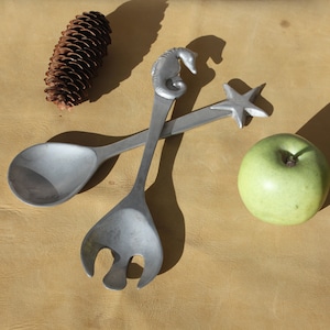 May include: Two silver salad servers with seahorse and starfish designs, next to a green apple and a pine cone. The servers are made of a metallic material and are set against a tan background. The seahorse server is approximately 25 cm long.