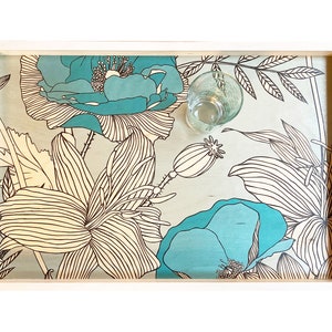 Puede incluir: Una bandeja de madera blanca con un diseño floral azul y blanco. La bandeja tiene una taza de vidrio en el centro.