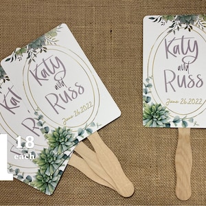 May include: Set of four white wedding fans with green succulent and eucalyptus illustrations. The fans feature gold lettering with the names "Katy and Russ" and the date "June 26, 2022".