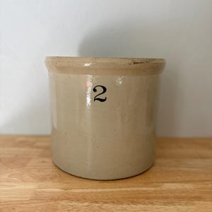 May include: A cylindrical stoneware crock with a smooth, light beige glazed finish. The exterior features a prominent black numeral 2 printed on the front. The rim has a slightly thicker, unglazed band with a visible chip on the upper edge.