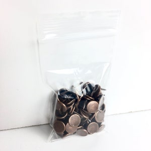 Puede incluir: Una bolsa de plástico transparente llena de una gran cantidad de pequeñas tachuelas redondas de color cobre.