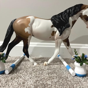 Pode incluir: Um modelo de cavalo com pelagem malhada castanha e branca, crina e cauda pretas. O cavalo está entre duas varas de salto brancas e azuis, cada uma ladeada por um pequeno vaso branco com flores artificiais azuis e creme no interior.