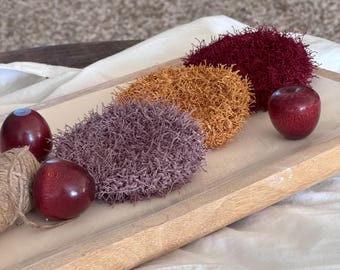 Op de afbeelding: Drie ronde, getextureerde schuursponsjes in de kleuren mauve, mosterdgeel en donkerbordeaux, gerangschikt op een rechthoekig houten dienblad samen met drie kleine, gladde, donkerrode decoratieve bollen en een klein bolletje jutegaren.