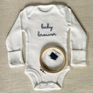 Op de afbeelding: Witte baby romper met lange mouwen met de tekst "baby brouwer" geborduurd in blauwe draad. Een houten borduurring met blauwe draad is ook afgebeeld.