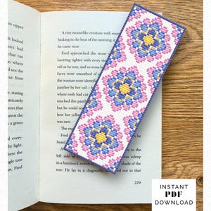 Puede incluir: Un marcapáginas rectangular de punto de cruz con un patrón floral repetitivo en tonos rosa, azul claro y amarillo sobre fondo blanco, con un borde cosido en azul oscuro, colocado sobre una página de libro abierta con texto impreso.