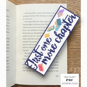 以下が含まれることがあります： 青いクロスステッチで「Just one more chapter」と書かれた手作りのブックマーク。ブックマークにはカラフルなステッチデザインがあり、開いた本の上に置かれています。