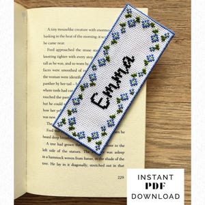 Könnte beinhalten: Ein personalisiertes, gesticktes Lesezeichen mit dem Namen "Emma" in schwarzen Buchstaben. Das weiße Lesezeichen ist mit blauen und grünen floralen Kreuzstichen umrandet. Das Lesezeichen liegt auf den Seiten eines Buches.