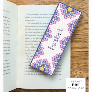 Könnte beinhalten: Ein rechteckiges Kreuzstich-Lesezeichen mit weißem Hintergrund und einem sich wiederholenden Blumenmuster in Rosa-, Blau- und Gelbtönen. Der Name Isabel ist in violetter Schreibschrift in der Mitte eingestickt. Das Lesezeichen hat einen dunkelblauen Rand.