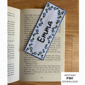 May include: A rectangular cross-stitch bookmark featuring a white fabric background with a border of small blue flowers and green leaves. The name Emma is stitched in black capital letters in the centre. The bookmark is placed on an open book page.