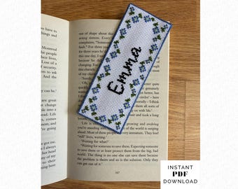 May include: A rectangular cross-stitch bookmark featuring a white fabric background with a border of small blue flowers and green leaves. The name Emma is stitched in black capital letters in the center. The bookmark is placed on an open book page.